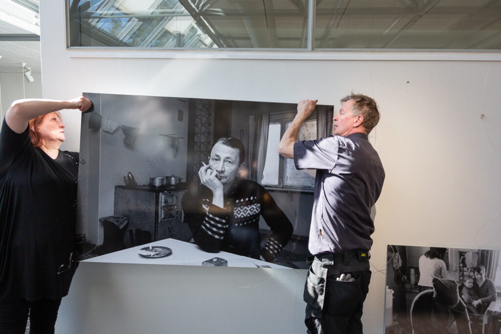 Opettaja Marjo Nurmi ja huoltomies Esa Sivusalo nostavat valokuvataulun siimojen varaan Murikka-opiston seinälle.