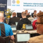 Useita ihmisiä istuu kokoussalissa. Puheenjohtajat ovat edessä kasvot kameraan päin. Muut istuvat kasvot poispäin kamerasta. Valkokankaalle on heijastettu kuva, jossa lukee "Teollisuusliiton sääntömääräinen kevätkokous".
