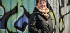 Nainen seisoo takki päällänsä ja huivi kaulassansa. Takana on seinä, johon on maalattu graffiteja.