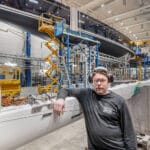 Seisaaltaan oleva mies nojaa palkkiin tehdashallissa. Miehellä on päässään kuulosuojaimet ja suojalasit.