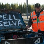 Huomioliiveihin pukeutunut mies seisoo auton vieressä. Auton konehuoneen kansi on auki. Kannessa lukee isoin kirjaimin "peli selvä".