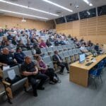 Kuva auditoriosta, jossa on kymmeniä ihmisiä. Useita penkkejä on myös tyhjänä.