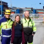 Kolme työntekijää seisoo tehdasalueen portin ulkopuolella.