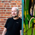 Mies seisoo ja katsoo kameraan. Hänen takanaan on tiiliseinä ja hän nojaa vieressä olevaan graffiteilla peitettyyn seinään.