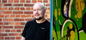Mies seisoo ja katsoo kameraan. Hänen takanaan on tiiliseinä ja hän nojaa vieressä olevaan graffiteilla peitettyyn seinään.