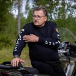 Mustiin pukeutunut mies nojaa moottoripyörään ja selittää kädellään viittoen. Takana on metsää.
