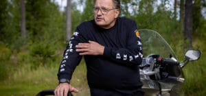 Mustiin pukeutunut mies nojaa moottoripyörään ja selittää kädellään viittoen. Takana on metsää.