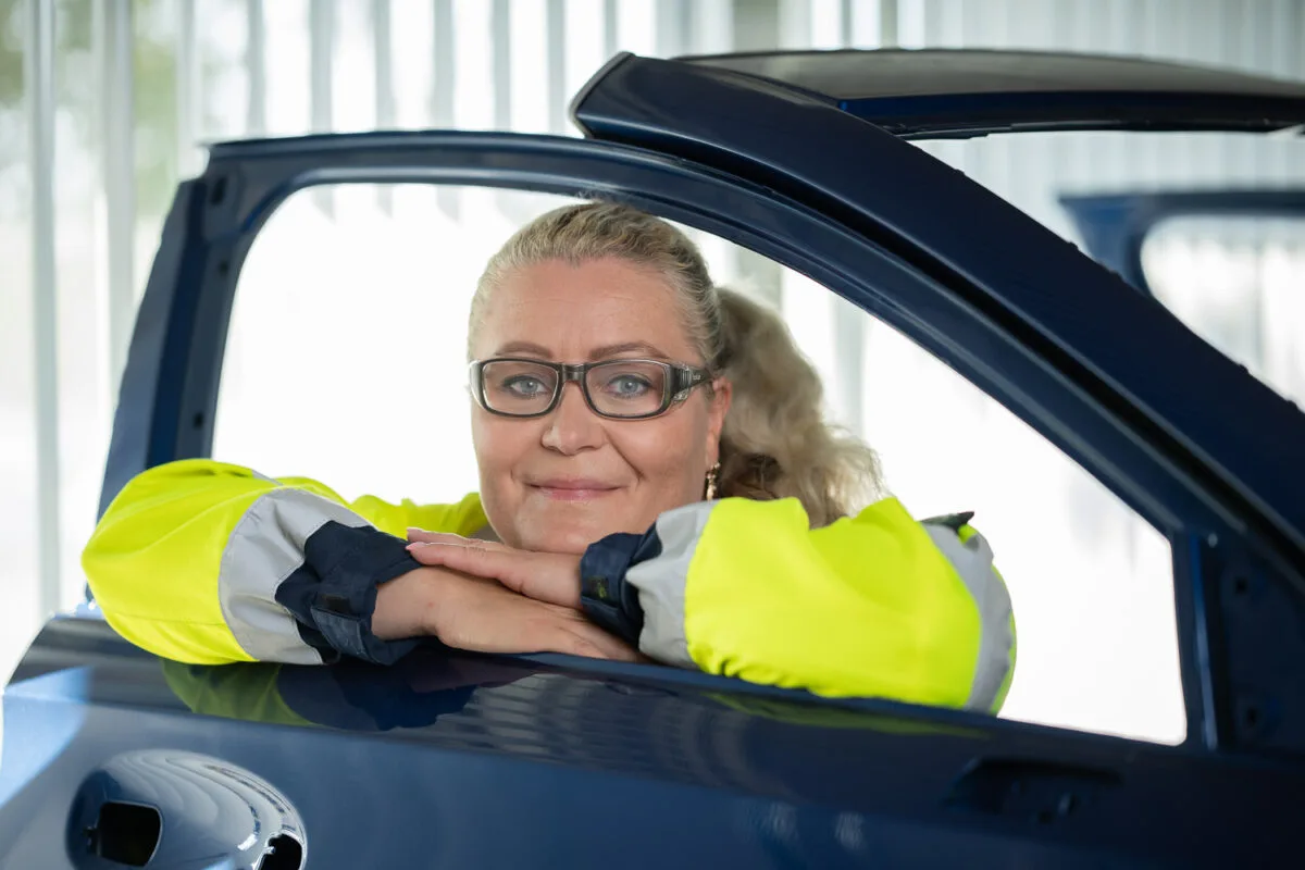 Nainen nojaa auton oveen ja katsoo avoimen sivuikkuna-aukon läpi. Hänellä on tehdastyöntekijän vaatteet päällään.