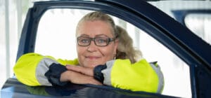 Nainen nojaa auton oveen ja katsoo avoimen sivuikkuna-aukon läpi. Hänellä on tehdastyöntekijän vaatteet päällään.