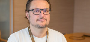 Henkilö katsoo kameraan puolilähikuvassa. Hän on sisätilassa ja hänellä on kaulassaan avainnauha, jossa lukee Teollisuusliitto.