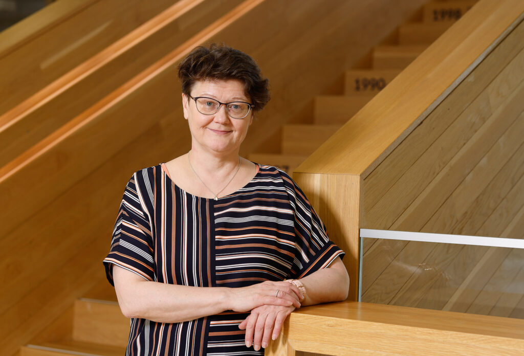 Sisällä kuvattu nainen nojaa puiseen kaiteeseen. Takana on puiset raput.