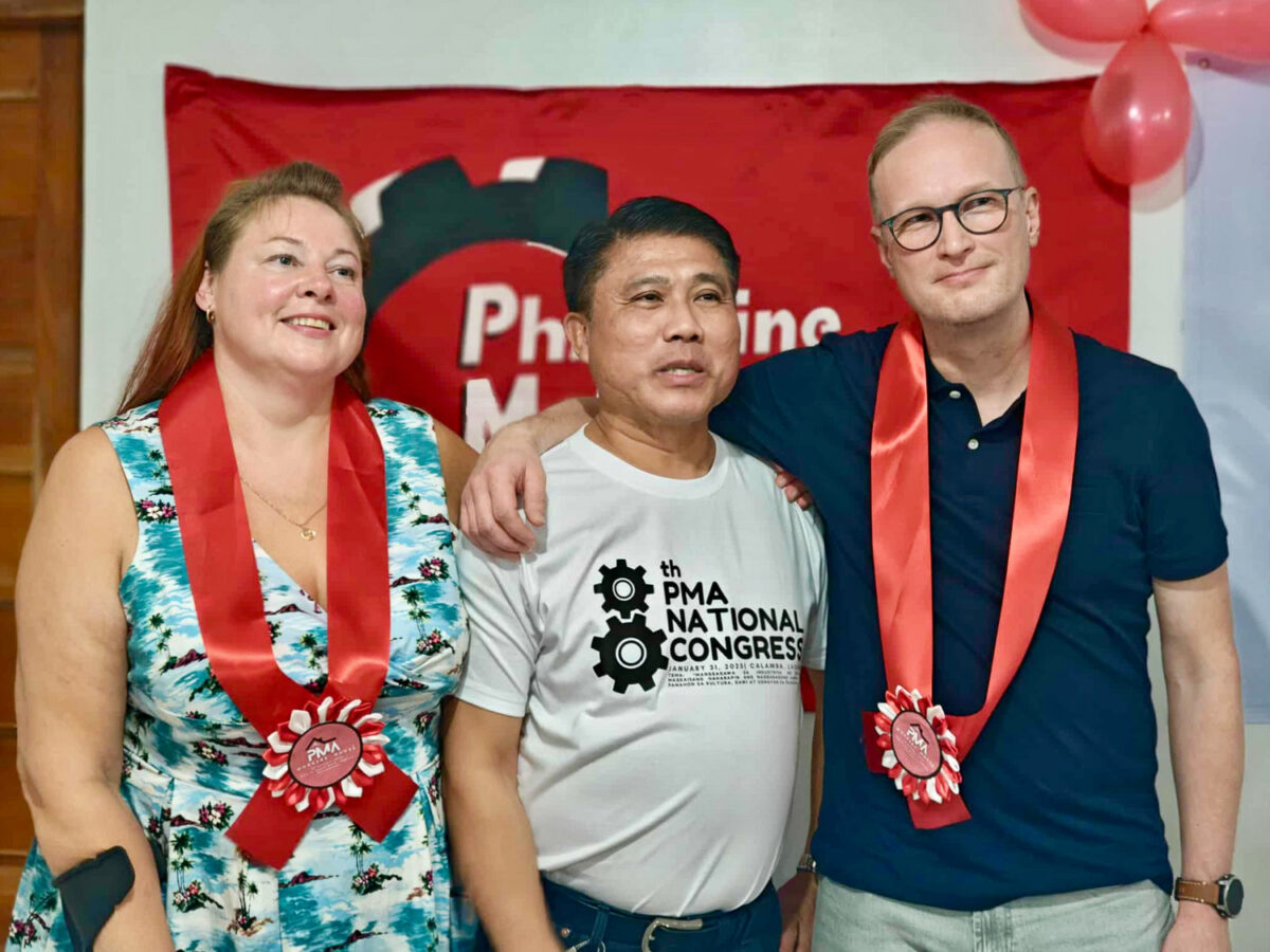 Kolme henkilöä seisoo rivissä kädet toistensa olkapäivällä. Takana on ammattiyhdistyksen punalippu.