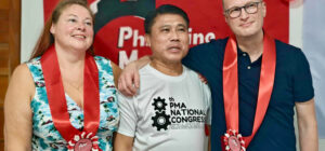 Kolme henkilöä seisoo rivissä kädet toistensa olkapäivällä. Takana on ammattiyhdistyksen punalippu.