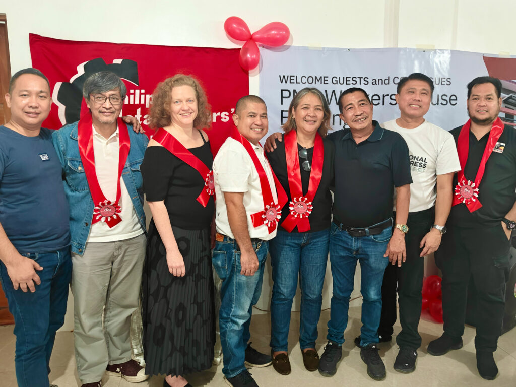 Yhdeksän henkilöä rivissa kädet toistensa olkapäillä. Takana on ammattiyhdistyksen lippuja.