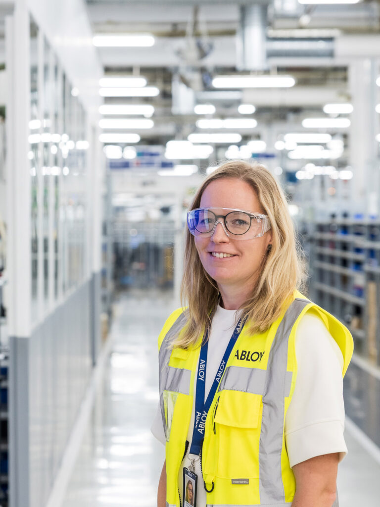 Henkilö seisoo tehdassalissa suojalasti päässä ja katsoo kameraan. Takana näkyy tehdassali ja kirkkaita valoja.