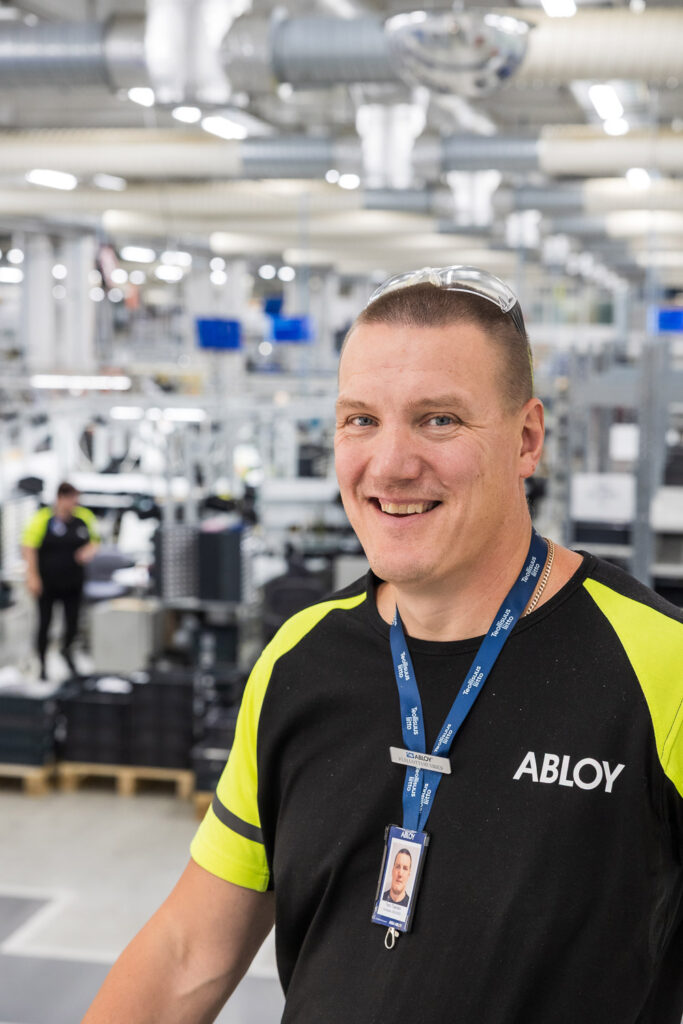 Henkilö hymyilee ja katsoo kameraan. Takana näkyy kirkasvaloinen tehdassali.