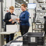 Kaksi henkilö seisoo tehdassalissa. He katsovat paperiliuskaa, jota he pitävät toinen toiselta puolelta kiinni.