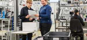 Kaksi henkilö seisoo tehdassalissa. He katsovat paperiliuskaa, jota he pitävät toinen toiselta puolelta kiinni.