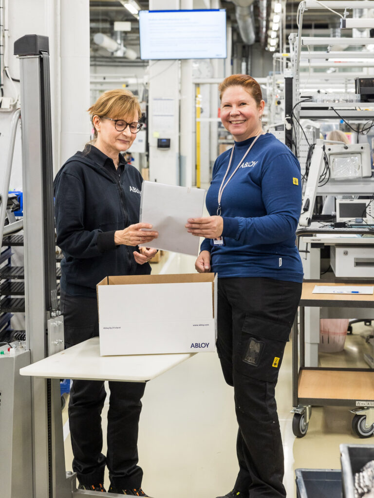 Kaksi henkilöä seisoo tehdassalissa. Heidän edessään tasolla on avoinainen pahvilaatikko.