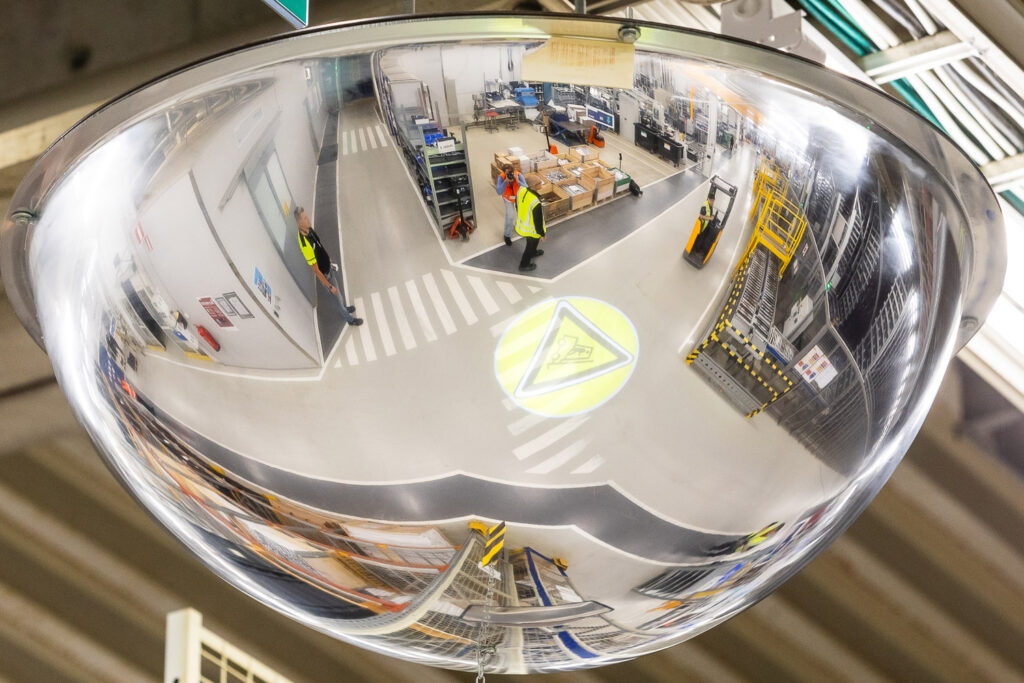 Tehdassalin kattoon kiinnitetystä puolipallon muotoisesta peilistä näkyy tehdassalin kulkuväyliä, työntekijöitä ja trukki.