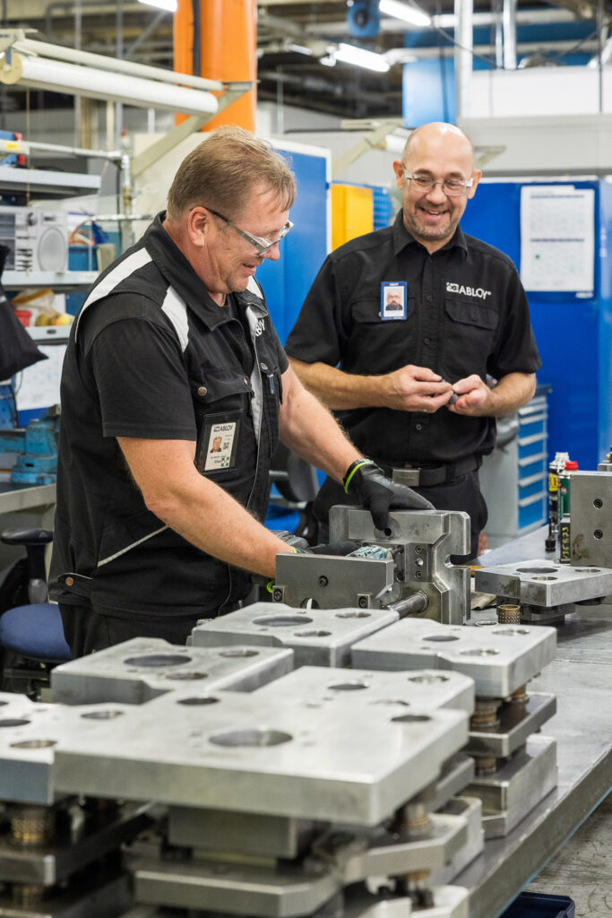 Kaksi työntekijää hymyilee tehdassalissa. Pöydällä heidän edessään on isoja metalliosia.