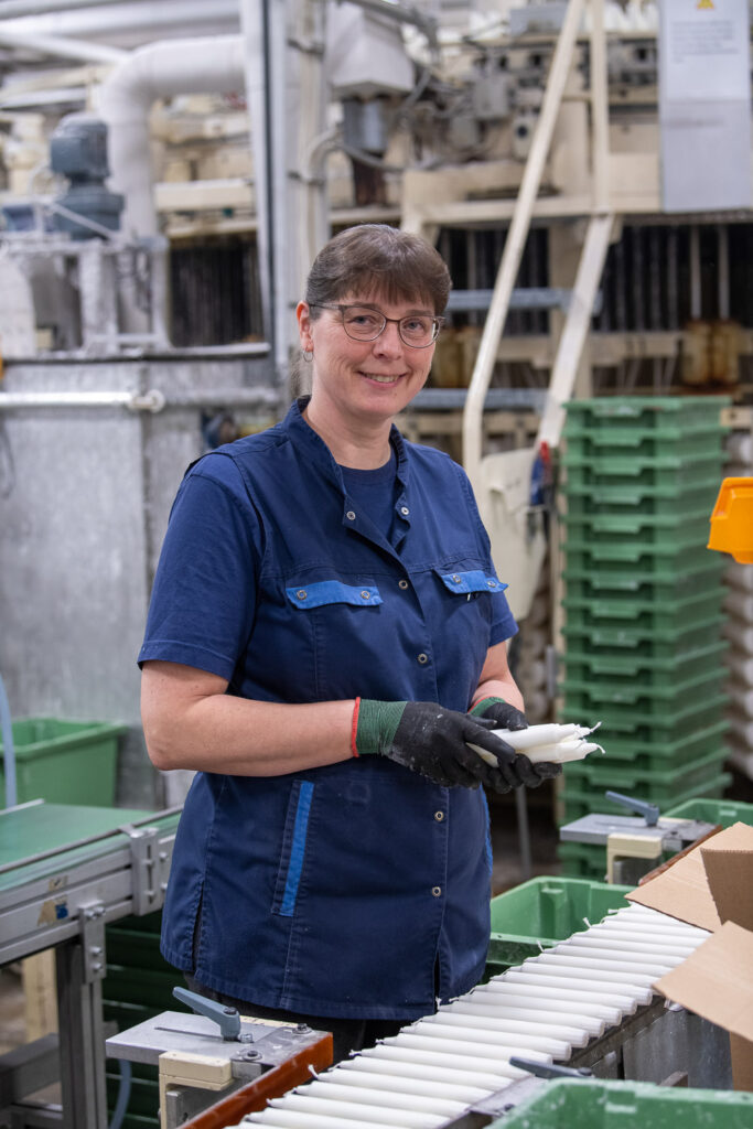 Henkilöllä on käsissä työhanskat ja hän pitelee kynttilänippua. Hän katsoo kameraan ja hänen edessään on kynttilärivi. Takana näkyy koneita ja rappuset.