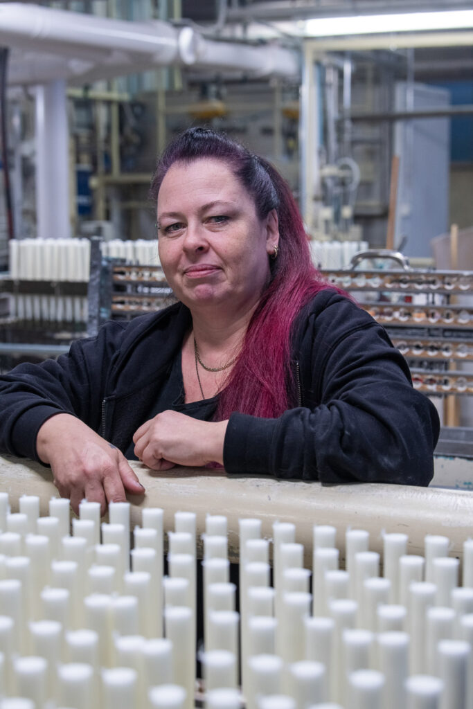 Henkilö seisoo kynttilälinjaston takana ja katsoo kameraan. Hänen edessään on paljon kynttilöitä ja takana näkyy tehtaan rakenteita ja koneita.