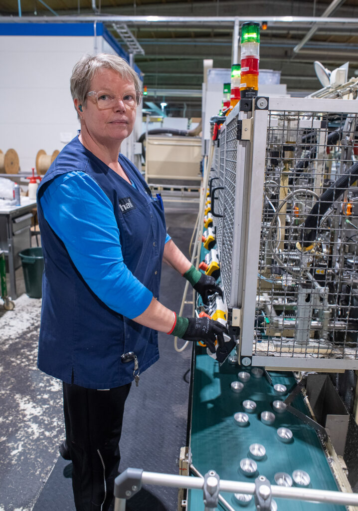 Henkilö seisoo tuikkukynttilöitä valmistavan koneen lähellä. Hänellä on kädet napailla, joita on pitkä rivi laitteen kyljessä. Tuikkukynttilöitä kulkee koneen alapuolella hihnan päällä.