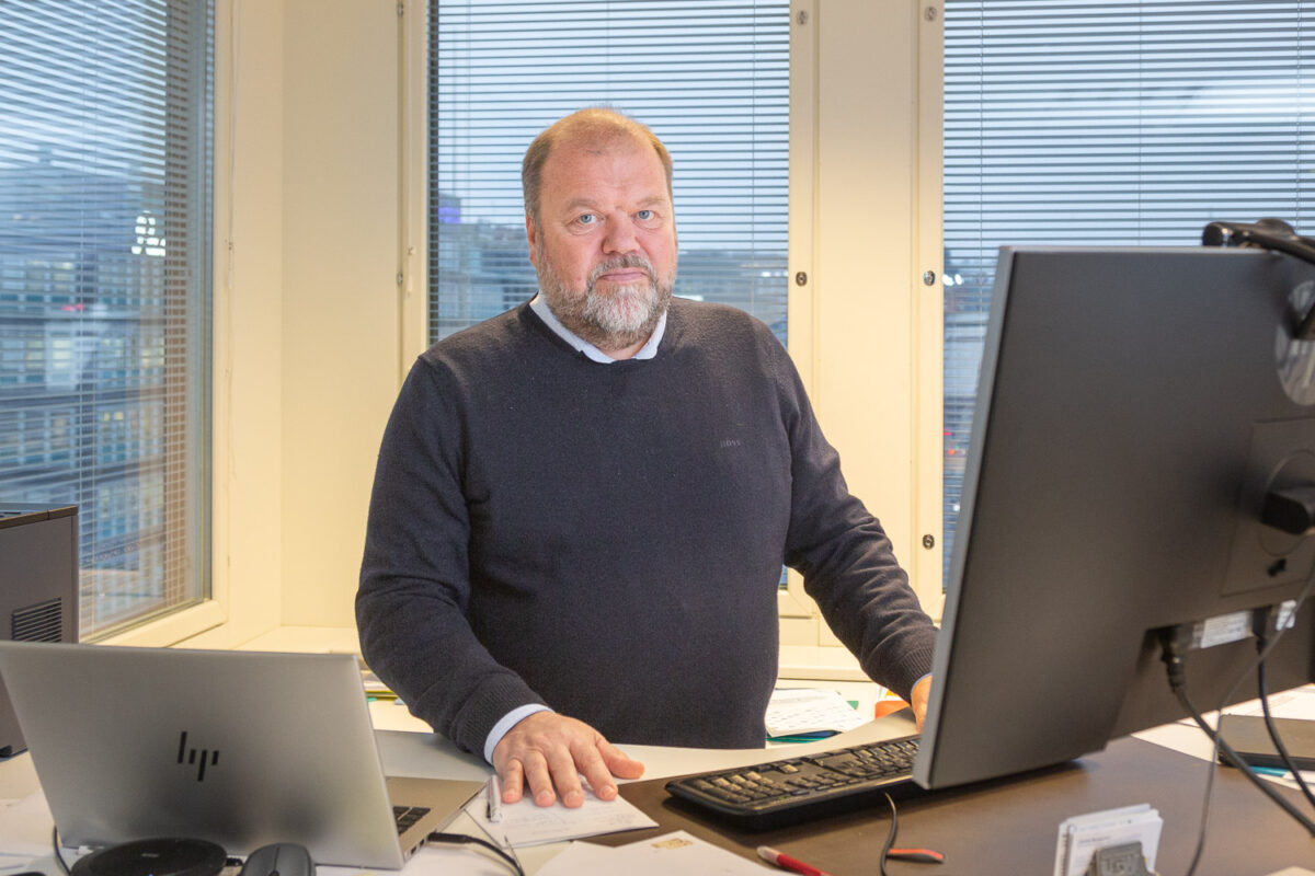 Henkilö seisoo työpöytänsä takana. Edessään hänellä on tietokone, ja takana ikkunoista näkyy harmaa kaupunkimaisema.