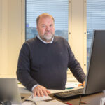 Henkilö seisoo työpöytänsä takana. Edessään hänellä on tietokone, ja takana ikkunoista näkyy harmaa kaupunkimaisema.