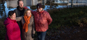 Neljä henkilöä seisoo tiiviissä ryhmässä metsässä ja heidän takanaan näkyy vesistö. Aurinko paistaa matalalta, ja vedestä heijastuu vastarannalla oleva valkoinen rakennus.