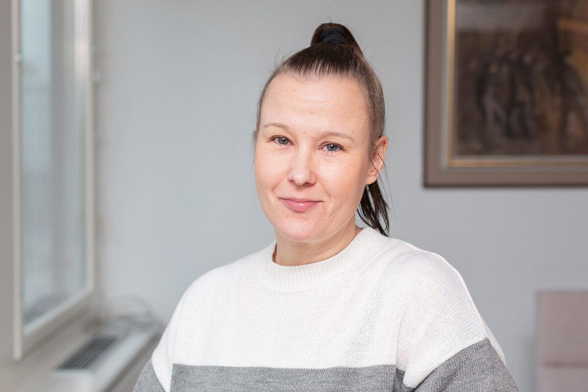 Blogin kirjoittaja Päivi Mäensivu on lähikuvassa sisätilassa. Hän katsoo kameraan. Takana näkyy valkea seinä ja taulun kulma.