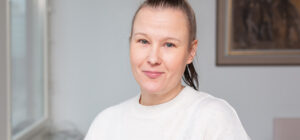 Blogin kirjoittaja Päivi Mäensivu on lähikuvassa sisätilassa. Hän katsoo kameraan. Takana näkyy valkea seinä ja taulun kulma.