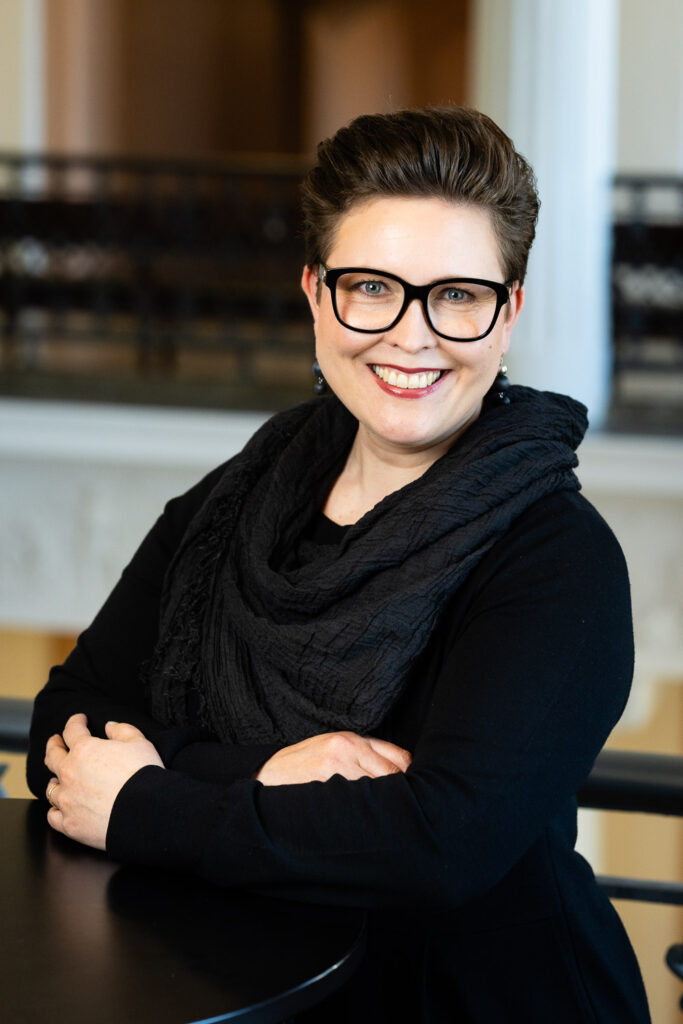 Sisätiloissa oleva henkilö katsoo kameraan, hän näkyy vyötäröstä ylöspäin. Takana nakyy kaide ja pylväs.