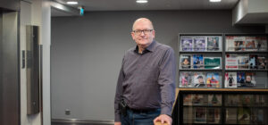 Sisätilassa seisova henkilö katsoo kameraan ja nojaa kädellä kaiteeseen. Takana näkyy aikakauslehtiteline ja hissin ovi.