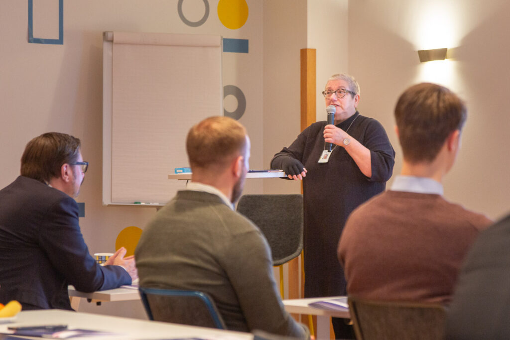 Nainen puhuu mikrofoniin. Kolme miestä kuuntelee. 