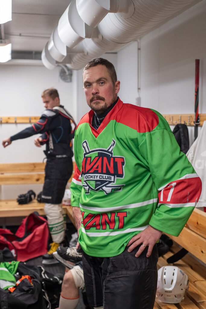 Mies seisoo pukuhuoneessa jääkiekkovarusteet päällään. Takana näkyy puupenkkejä ja toinen pelaaja.