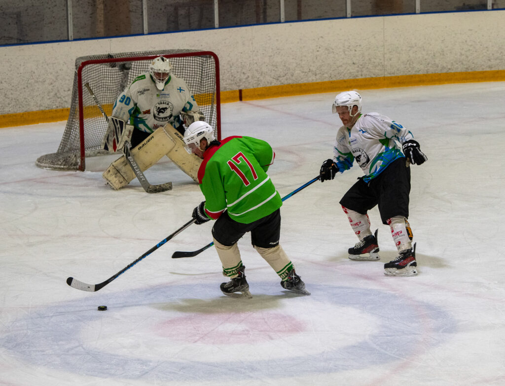 Jääkiekkoilijat pelaavat jäähallissa. Näkyvissä on hyökkääjä, jolla on kiekko, sekä toisen joukkueen puolustaja, maalivahti ja maali.
