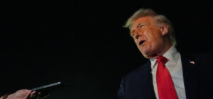 Sivusta kuvattu henkilö puhuu. Toisesta reunasta näkyy käsi, joka pitelee äänen tallennuslaitetta. Tausta on pimeän musta.