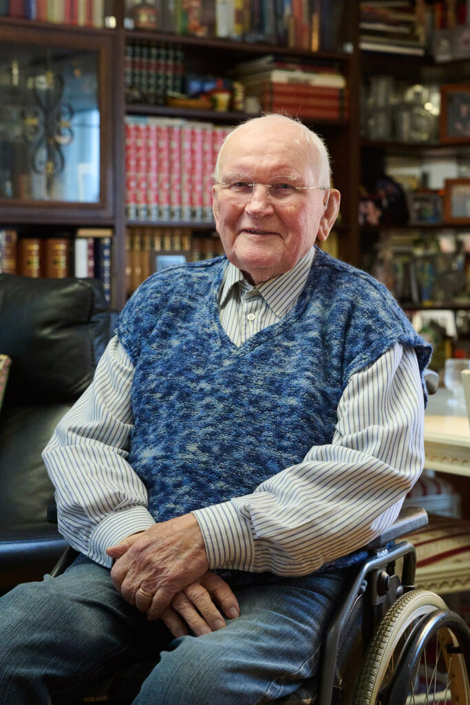 Henkilö istuu pyörätuolissa ja hymyilee. Takana näkyy kirjahylly.