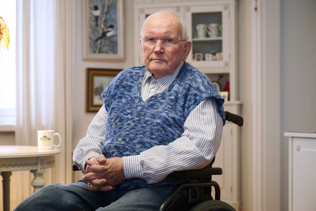 Henkilö istuu kädet ristissä pöyrätuolissa. Vieressä olevalla pöydällä on muki ja takana näkyy kaapissa lasioven takana olevia mukeja.