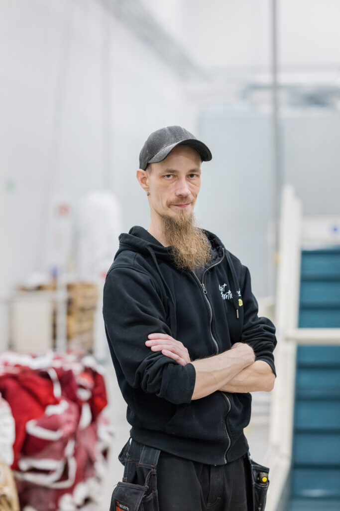 Henkilö seisoo kädet puuskassa ja katsoo kameraan. Taustalla näkyy tuotantolaitoksen sisätiloja.