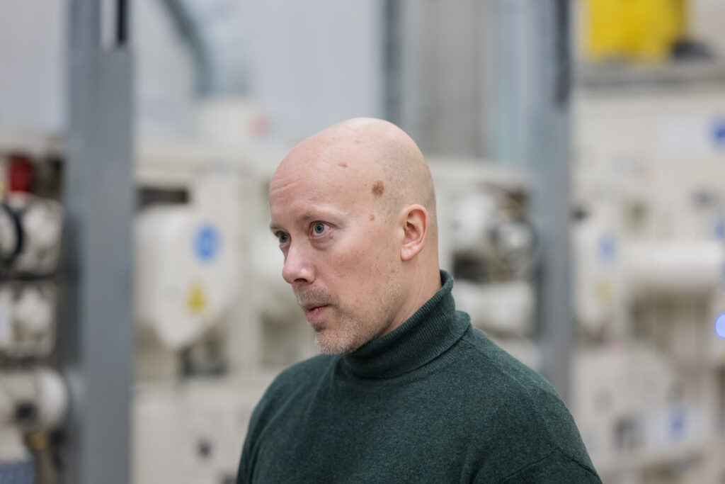 Läheltä kuvattu mies katsoo kameran ohi. Takana näkyy tuotantolaitteita.
