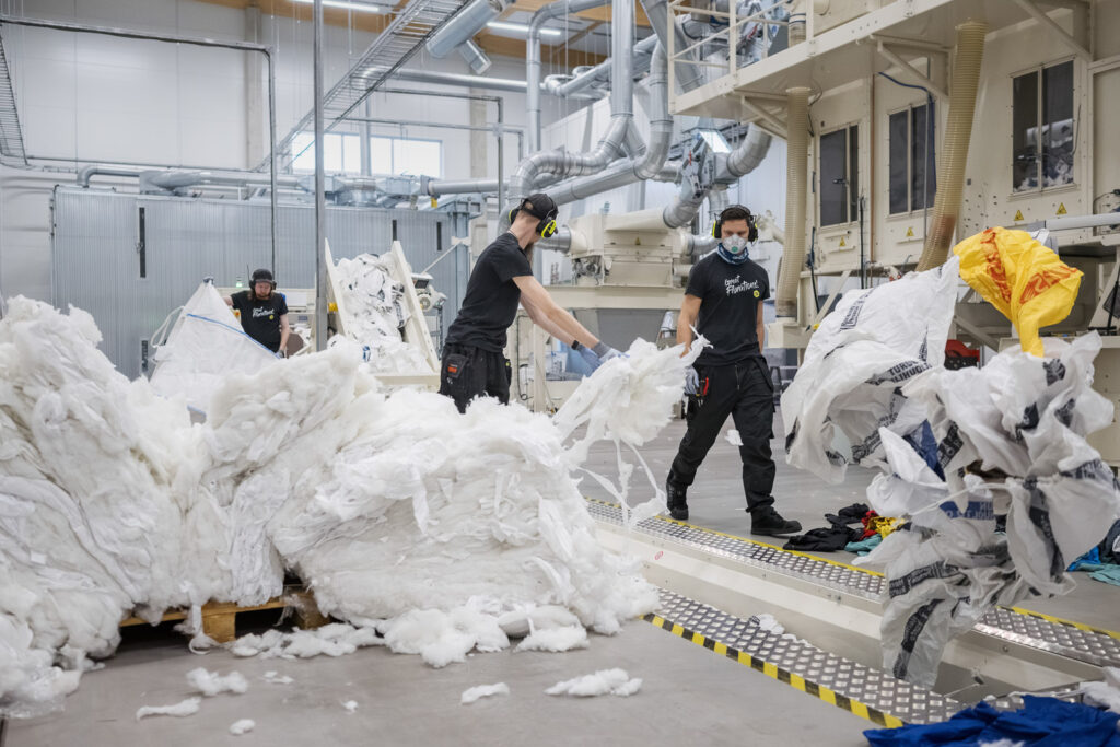 Kolme työntekijää siirtää kierrätettyä kangasmateriaalia tuotantolaitoksen prosessissa sisätilassa.