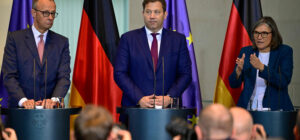 Kolme henkilöä seisovat yleisön edessä korokkeella. Heidän takanaan on sinisiä sekä musta-puna-keltaisia lippuja.