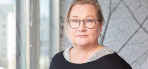 Sisätilassa läheltä kuvattu henkilö katsoo kameraan. Takana näkyy kuviollinen seinä ja ikkuna.