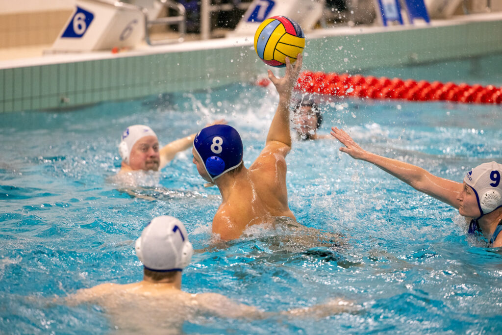 Viisi henkilöä pelaa vesipalloa uima-altaassa. Hyökkäävä pelaaja valmistautuu heittämään palloa.