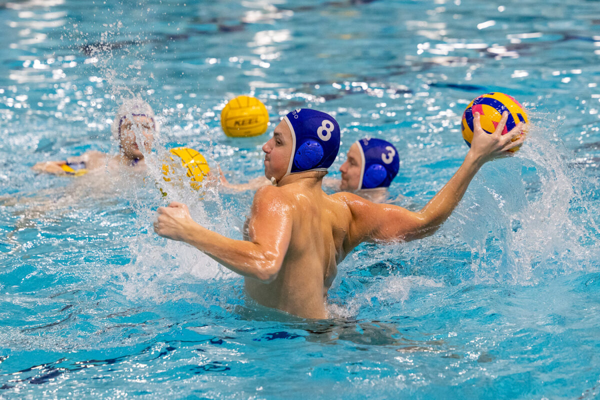 Vesipalloa pelaavia henkilöitä uima-altaassa. Yksi pelaajista ponnistaa vedestä ylös ja valmistautuu heittämään palloa.