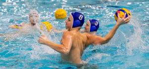 Vesipalloa pelaavia henkilöitä uima-altaassa. Yksi pelaajista ponnistaa vedestä ylös ja valmistautuu heittämään palloa.