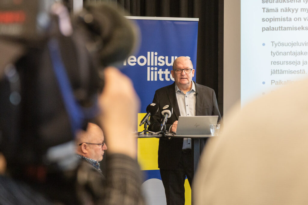 Mies puhuu neuvotteluhuoneessa. Etualalla kamera.